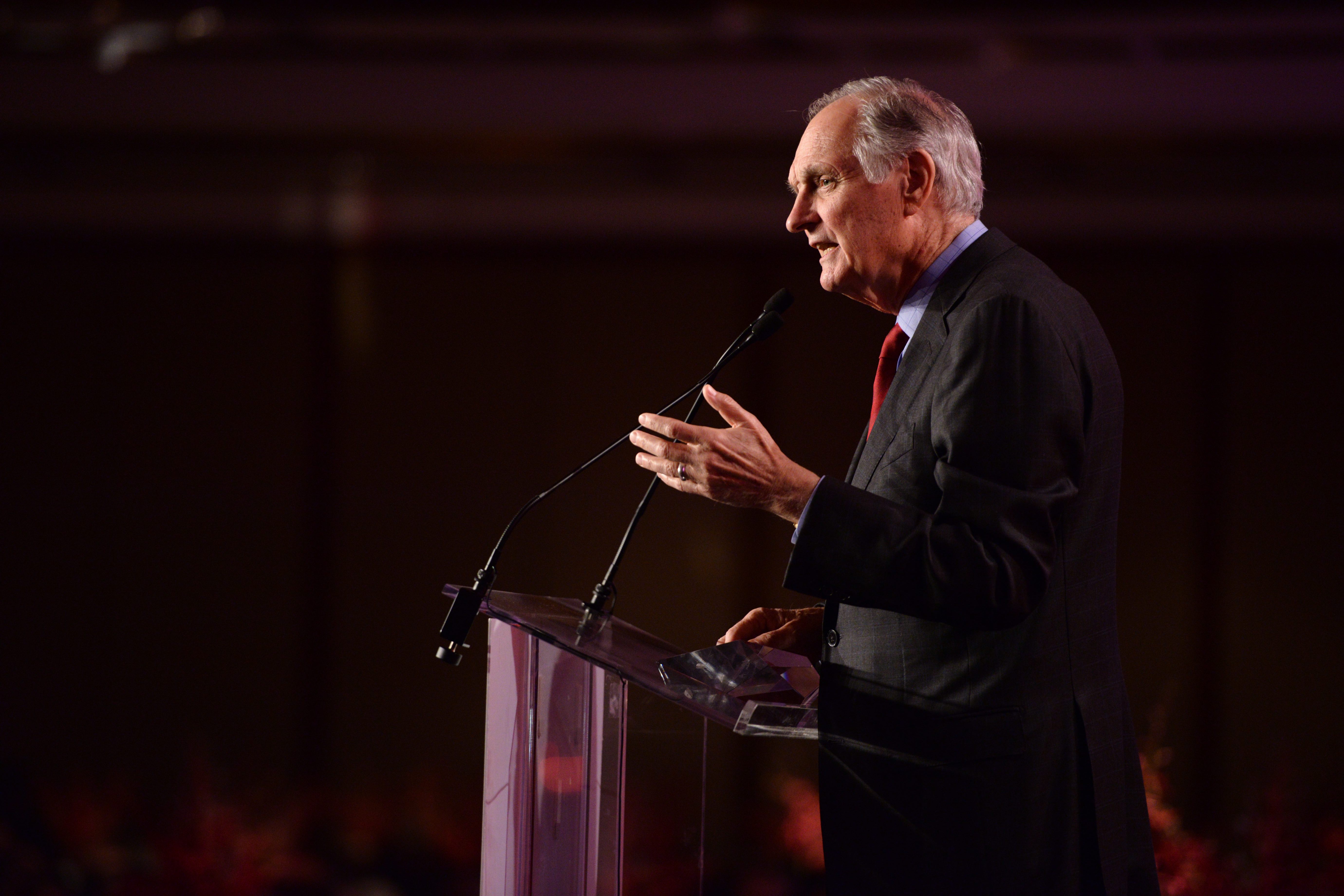 Alan Alda stands at a lectern on a stage in profile giving a talk