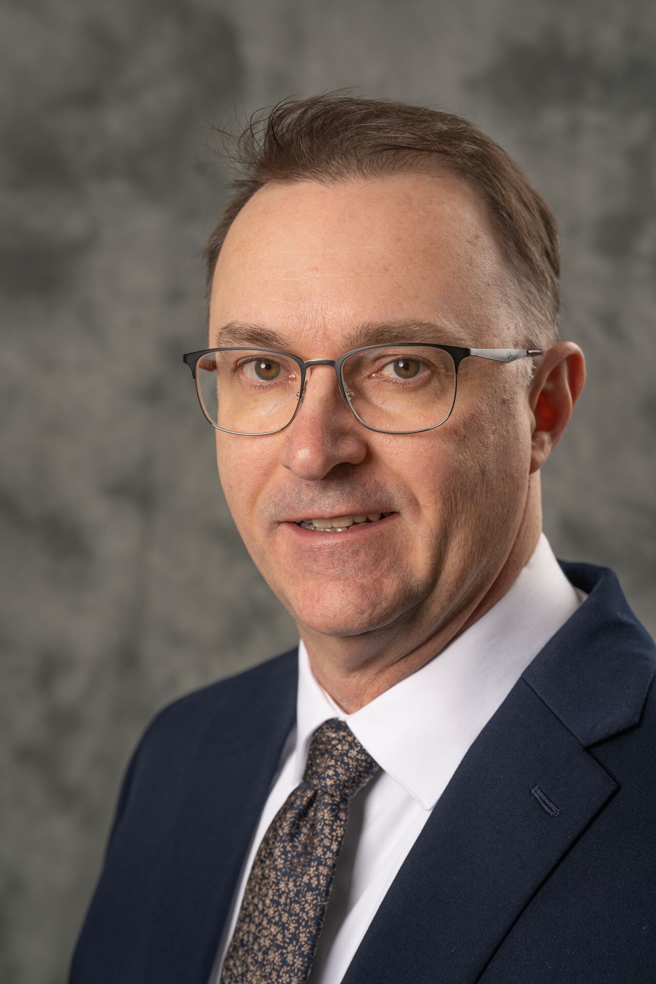 Portrait of a professional in a dark blue suit and glasses, against a mottled gray background.