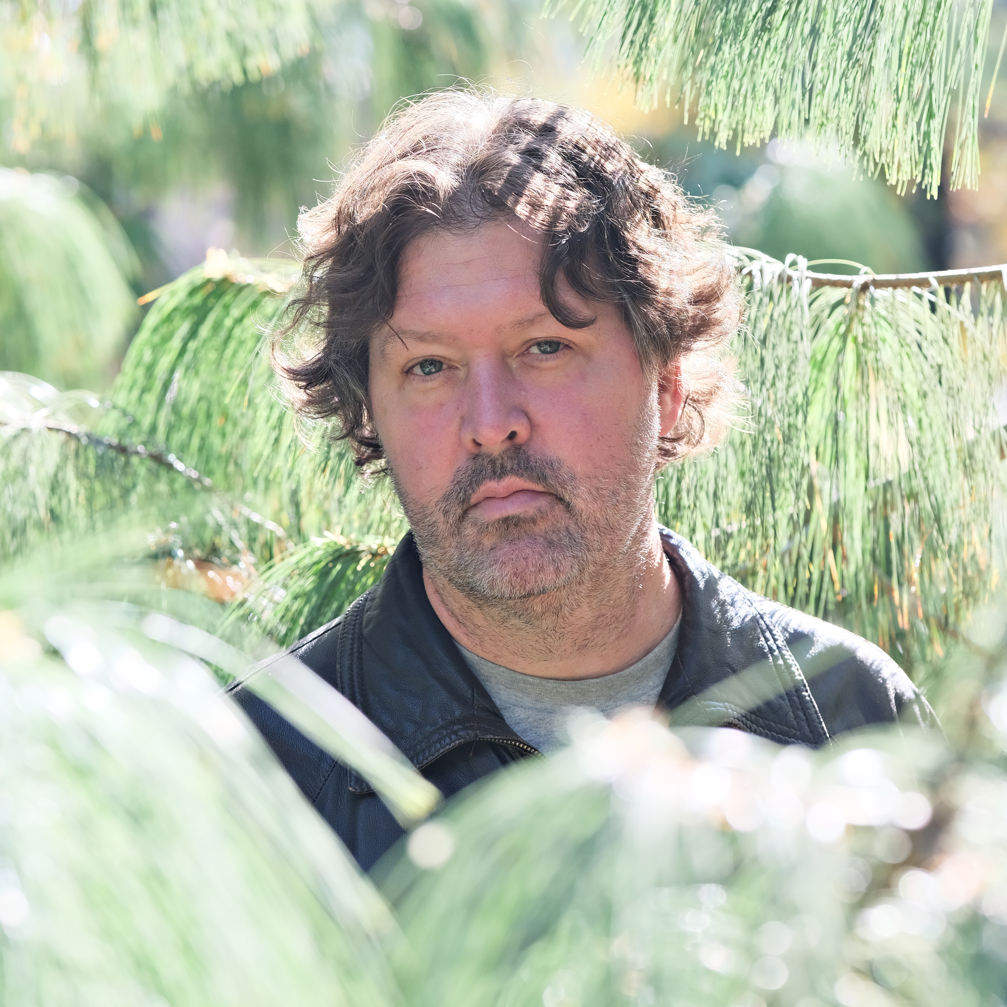 person with brown wavy hair standing in front of a green tree