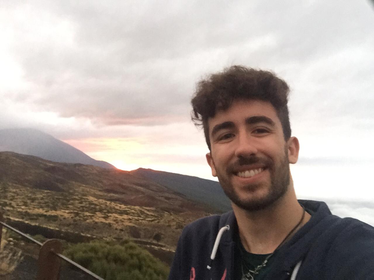 A person smiling at the camera with a beautiful sunset and mountains in the background.