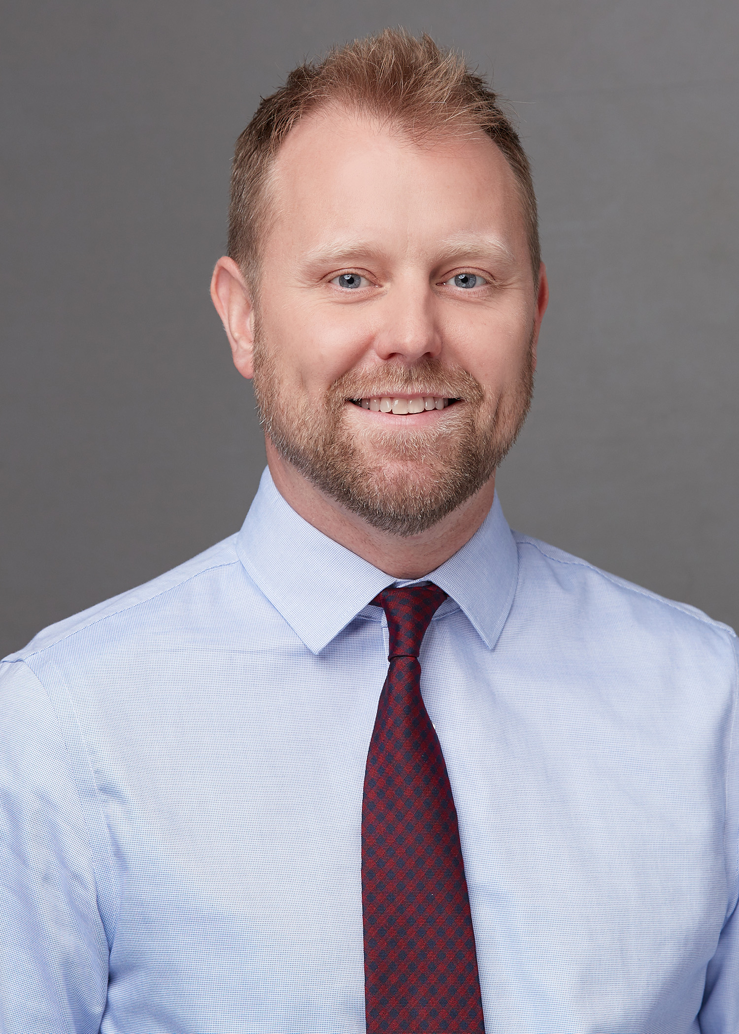 A professional portrait of a smiling person wearing a light blue shirt and a checkered tie against a grey background.