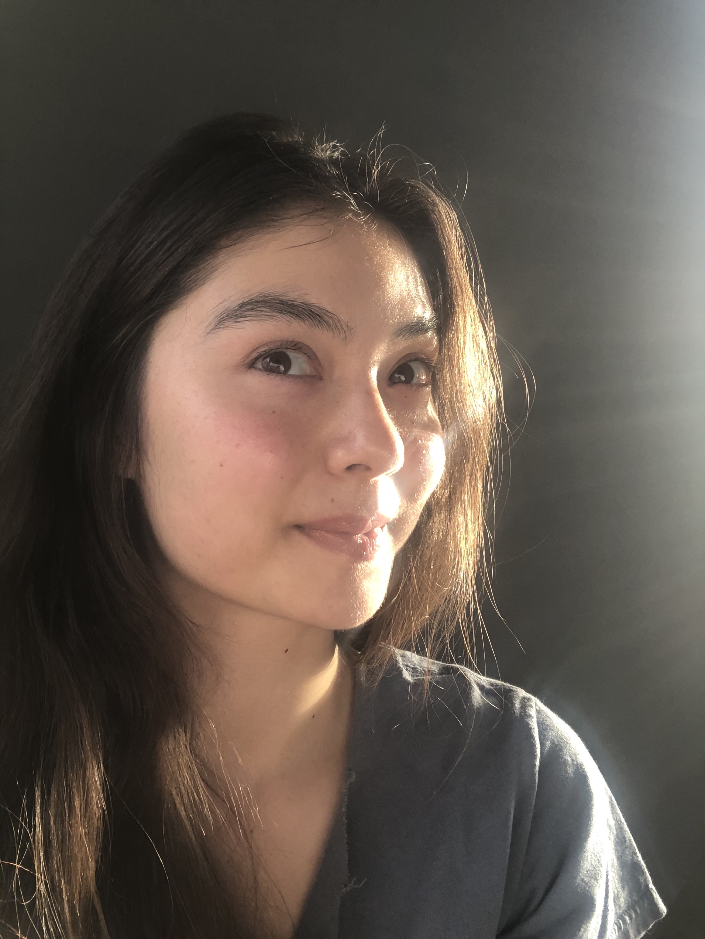 Close-up portrait of a person with long hair, gazing slightly away from the camera, with sunlight illuminating one side of their face.